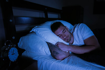 Handsome man asleep in bed at night with traditional alarm clock on the bedside table next to him.  