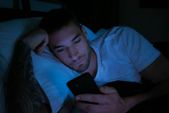 Handsome man looking at his mobile phone in bed at night with glow of phone lighting his face