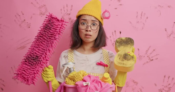 Domestic cleaning for you. Unhappy woman looks puzzled feels dissatisfied as did housework all day long gazes at mop and dirty spone provides household service wears hat with brush rubber gloves