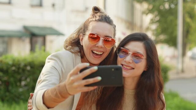 Positive emotions and love. Two young smiling hipster blond women taking selfie self portrait photos on smartphone. Pretty stylish girls on street background. LGBTQI, Pride Event, LGBT Pride Month