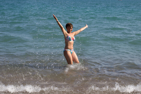 Schlanke Frau mit kurzen, dunklen Haaren und mit dem Bikini, spritzt mit dem Wasser im Meer. Sie lacht und hat Spaß im Wasser. Die Hände sind oben. Man sieht den ganzen Körper. Türkei, Fame Residence