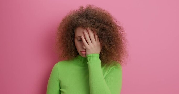 Serious curly haired young woman has uninterested bored expression shakes head makes face palm wears green turtleneck cannot listen dull beggar poses against pink bacground dressed casually.