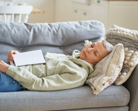 Senior grey haired woman sleeping on coach after reading book, having a nap while spending leisure time alone at home. Selective focus on senior female