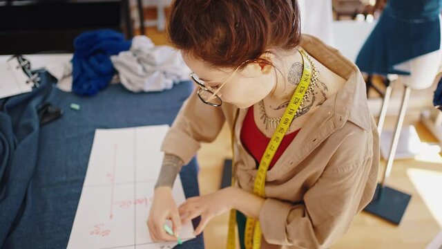 Over shoulder view of young woman seamstress preparing pattern for clothes, drawing it on fabric with chalk