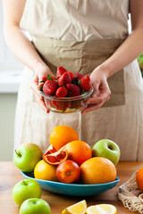Beautiful young woman holding bowl with ripe red strawberries. Big blue plate with apples, oranges on wooden table. Concept of healthy food, clean eating, low calories dessert, dieting. Close up