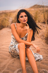 Caucasian brunette stroking her hair in a white floral dress, sitting on the sand on the beach of Cabo de Gata, Nijar. Andalucia, Spain