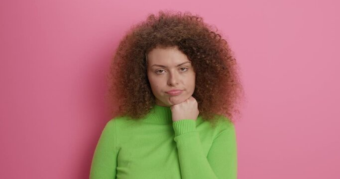 Indifferent beautiful curly haired woman shakes head keeps fist under chin cannot listen stupid talks anymore has bored face expression wears casual green turtleneck isolated over pink background
