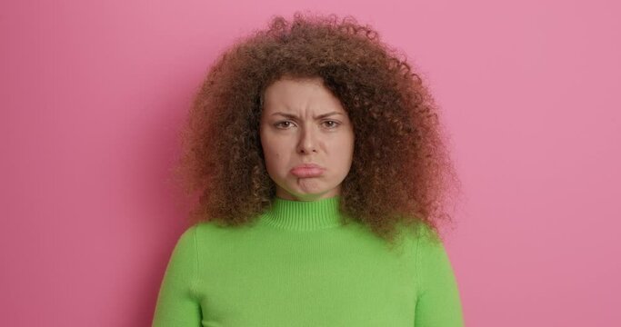 Surprised curly haired young woman covers mouth from wonder hears shocking bad news sulks face looks unhappily being worried about something wears casual clothes poses indoor. Human reactions