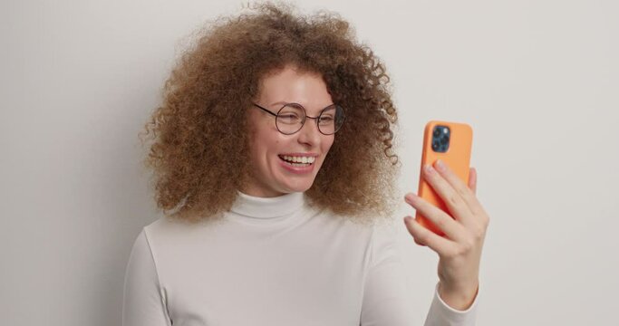 Overjoyed glad European woman with curly hair waves hello in smartphone greets someone feels very happy wears casual turtleneck isolated over white background enjoys friendly videoconference