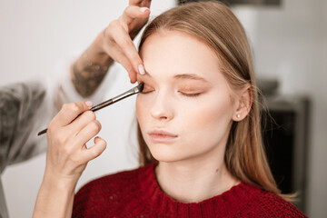 makeup artist applies eye shadow to the eyelids on a beautiful young woman blonde model, face make up concept