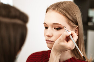 makeup artist applies eyebrow shadow on a beautiful young woman blonde model, face make up concept