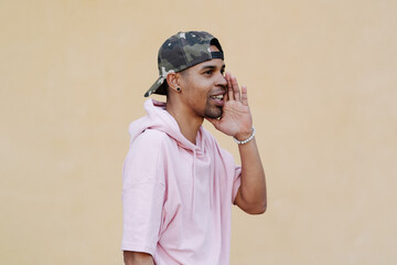 Attention. Close up portrait of cheerful young afro man in pink parka is holding hand near his open mouth isolated on pale yellow background with copy space for text