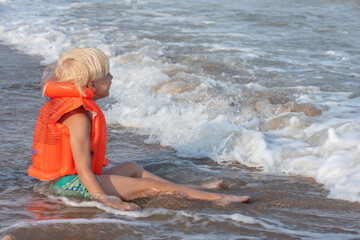 Blond boy in an inflatable swimming vest sits on seashore in the water.