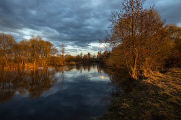 Rural landscape with a river. Trees, bushes and the ground are illuminated by the rays of the setting sun. The sky is covered with clouds. Lead clouds are reflected in the water.