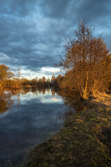 Rural landscape with a river.Trees, bushes and the ground are illuminated by the rays of the setting sun.The sky is covered with clouds.Lead clouds are reflected in the water.A center on the horizon
