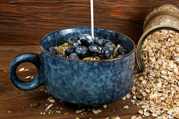 Pouring milk in an oatmeal porridge with blueberries and seeds in a blue ceramic bowl on a wooden background.
