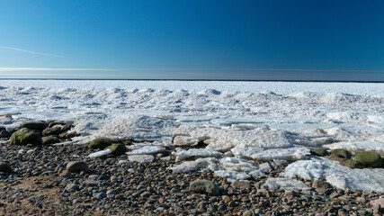 winter landscape by the sea, ice and stones in the dune area, in the distance the sea horizon, blue sky