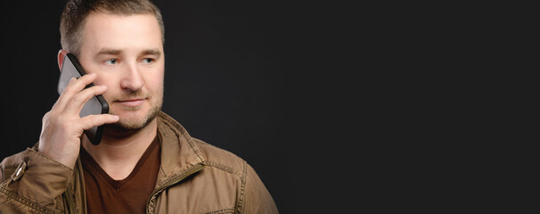 Panoramic attractive positive Caucasian man in casual clothes is talking on the cellphone and looking to the side. Studio shot on a black background. Phone call delivery and service concept