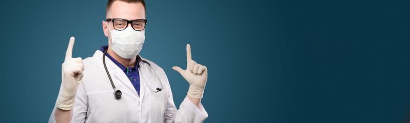 panoramic Attractive young Caucasian male doctor in protective mask and glasses shows two index fingers upwards and smiles. Studio shot on blue background