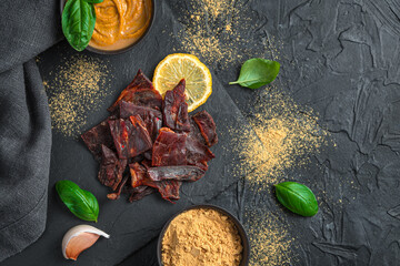 Slices of dried meat with garlic, fresh basil and lemon on a black background with space to copy.