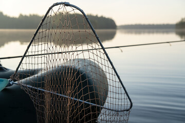 Net for pulling fish out of the water and spinning rods in an inflatable boat, at dawn, against the backdrop of a lake and forest in fog.