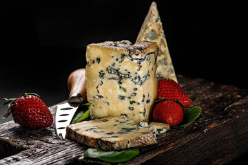 Semi-soft cheese with mold made from cows milk. The king of blue cheeses is Stilton. A piece of cheese and ripe strawberries on a dark wooden board and a cheese knife. Low key image. Copy space