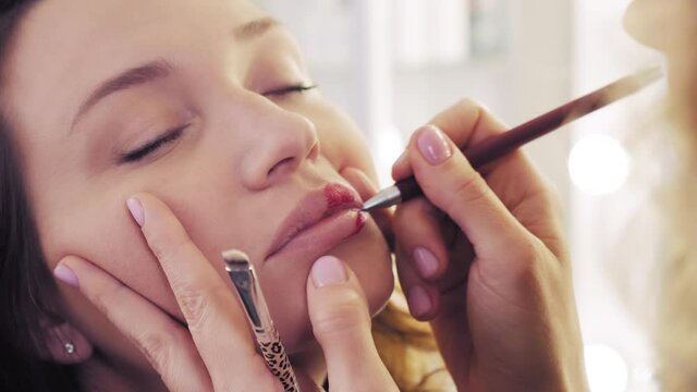 Make up artist applying lipstick. Closeup of the lips of a young woman, professional makeup artist with a brush applies red color lipstick. fashion backstage. Beauty salon.