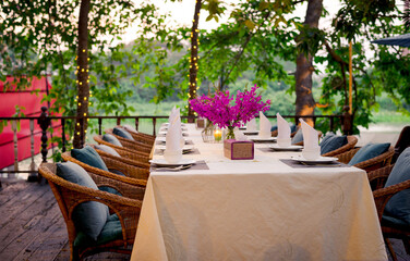 Vases with bouquets purple flowers, plates, tissue, napkin sets on the dinning table composite of rattan arm chairs and cushions with bushes flowers and small light lamp in the background.