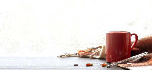 red mug and woolen blanket on a table with a thermometer and pills against the backdrop of a frosty...