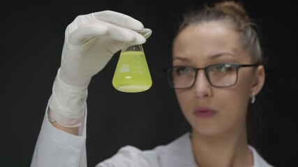 Young Caucasian Female Research Scientist Checking in Test flask yellow Chemical Liquid with dark precipitate at bottom. Microbiologists Conducting Biotechnology Research on black backdrop close up