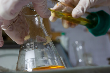 Gloved hands pour liquid from one flask into another, against a blurred background. Scientist is conducting an experiment in the laboratory. 