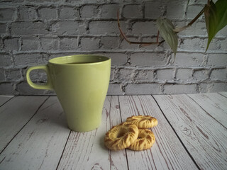 yellow mug with coffee, yellow cookies in the foreground, behind a white brick wall and a green plant