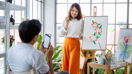 Obraz premium Senior woman artist painting image in studio at home with green small tree in little garden and full of natural light. Her older husband using smartphone to take her photo with love and care.