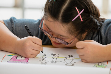 asian girl with down syndrome wearing eyeglasses sitting and use a color pencil drawing a family picture on a white paper willingly at home. Happy family concept