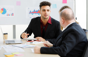 2 Adults caucasian businessmen are talking. A focused businessman is holding pen and look at a teammate and have brown cup, book, laptop put on desk and whiteboard are back with more graph on paper