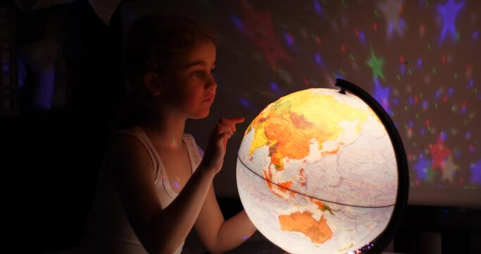 Girl Playing With Globe at Night. Child Sits on Bed in Evening Light Dreaming Vacation. Little Child Looking at Illuminated Globe, Exploring World, Learning. Dreaming About Future Save of Our Planet.