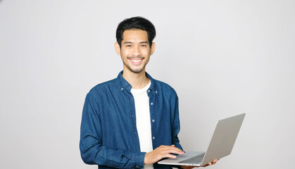 Young asian man holding laptop computer smiling and looking at camera standing on isolated grey...