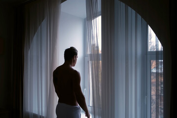 Fototapeta na wymiar Silhouette of young man in white towel in hotel near window in morning in city, side view. Traveling, resting, tourism concept. The hotel is located a historic building.