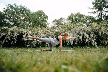 Young red-haired woman performs exercises for the spine and posture in the park. Lotus position in...