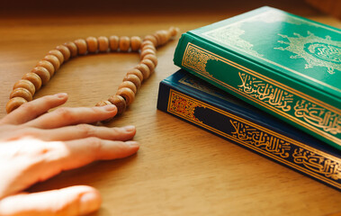 The holy  Quran with written arabic calligraphy meaning  "Holy Quran" and a rosary (beads) on the table, the hand of a man preparing for prayer during Ramadan