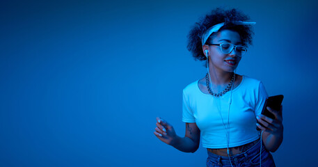 Trendy girl with afro curls listens and enjoys music with headphones in blue neon light, hip hop...