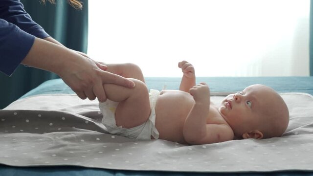 Mother with baby boy doing exercise simple exercises to make baby bones and muscles stronger, newborn infant lying on bed against the window. High quality 4k footage