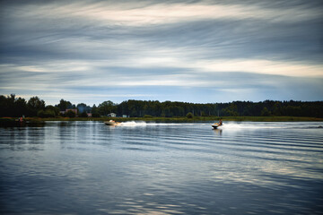 А jet ski and a boat quickly float along the calm calm river water with cloudy weather.