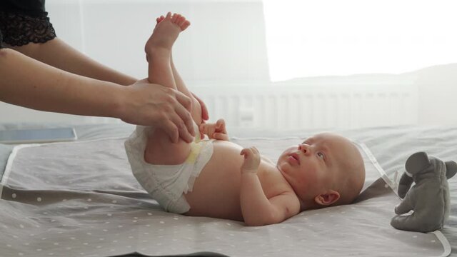 Mother with baby boy doing exercise simple exercises to make baby bones and muscles stronger, newborn infant lying on bed against the window. High quality 4k footage