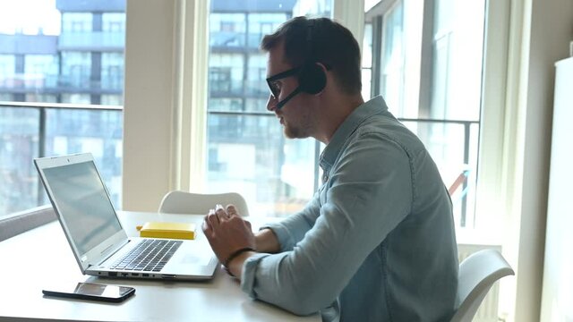 Side view at young male employee wearing headset talking online with a colleagues or customers, a man wearing casual shirt using hands free device for video communication on the distance
