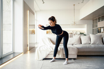 Obraz premium A brunette woman does a workout in a bright apartment. Body stretching exercises for the cardio system. Healthy lifestyle. Stylish sports black suit is tight-fitting.