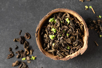 green dry tea leaf with sausep fruit in a coconut bowl with scattered leaves, gray background, close-up, top view