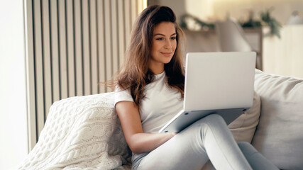 Fototapeta premium Young woman of European appearance online training on a laptop. Blogger housewife brunette sitting at home. Makes online purchases on the computer.