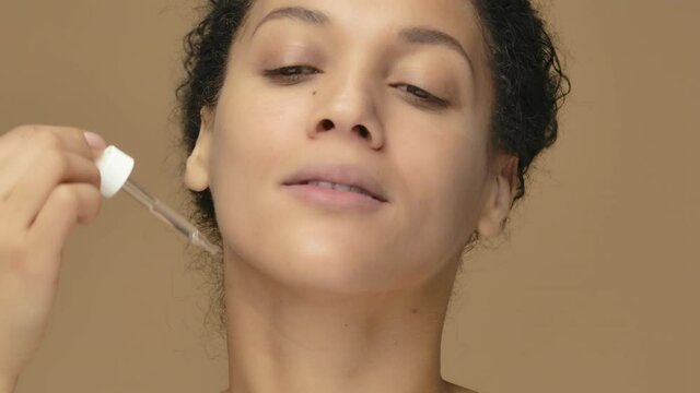 Beauty portrait young African American woman applying skincare serum on face, rubbing gently with fingers. Black female posing on brown studio background. Close up. Slow motion ready, 4K at 59.94fps.