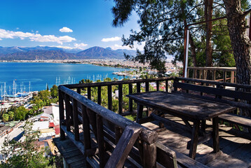 Obraz premium View of Fethiye harbor with yachts and boats and mountains. There is a wooden table with benches for resting in the foreground. Turkey, Mugla Province. 